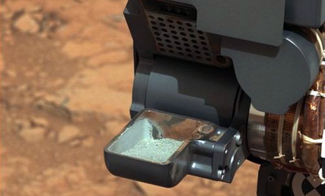 The Curiosity rover holding a scoop of powdered rock on Mars. The rover recently drilled into a Martian rock for the first time and transferred a pinch of powder to its instruments to analyze the chemical makeup.