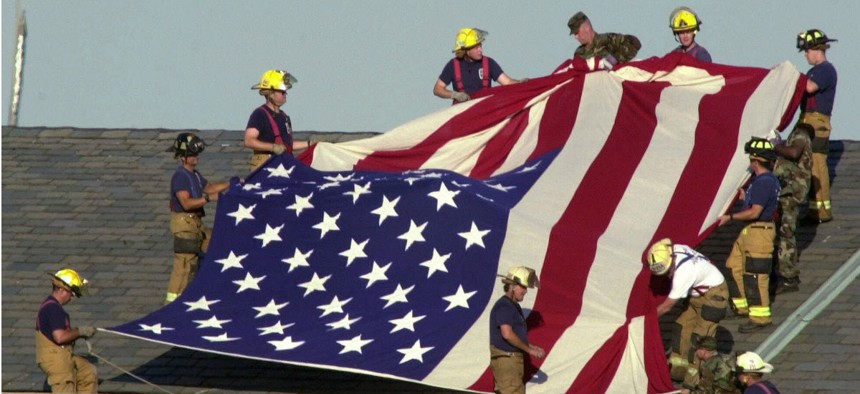 Military and fire personnel unfurl a flag on the roof of the Pentagon on Sept. 12, 2001. 