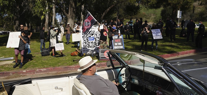 A driver in his convertible cruises past a small group of anti-COVID-19 vaccine protesters demonstrating at Elysian Park, outside the Dodger Stadium vaccination mass center in Los Angeles in February