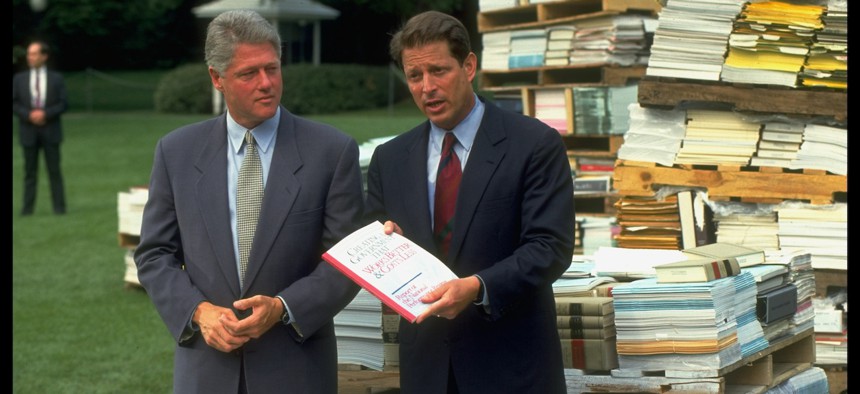 President Bill Clinton looks on as Vice President Al Gore presents his National Performance Review. The two are standing among piles of government regulations.