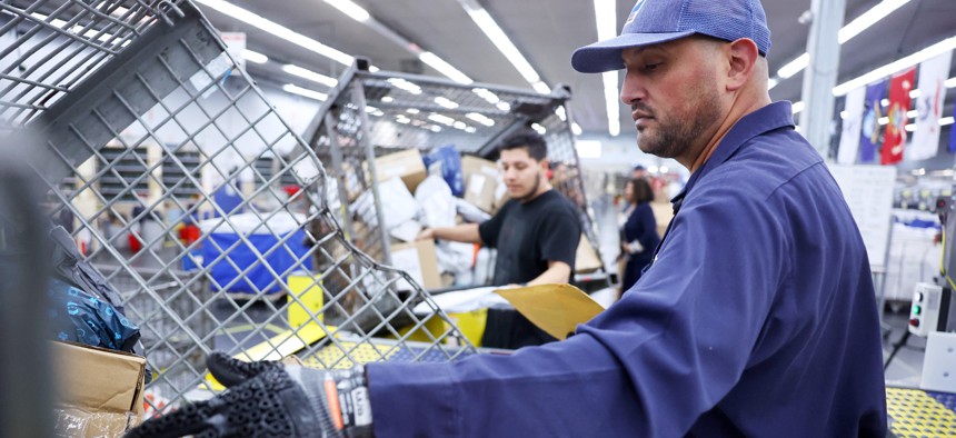 U.S. Postal Service employees Cesar Chavez, right, and Gilbert Gonzalez place packages into a new package sorting machine ahead of the holiday mail rush in Torrance, California, on Nov. 27, 2023. USPS employees, annuitants and eligible family members will enroll in a new health insurance exchange this fall.   