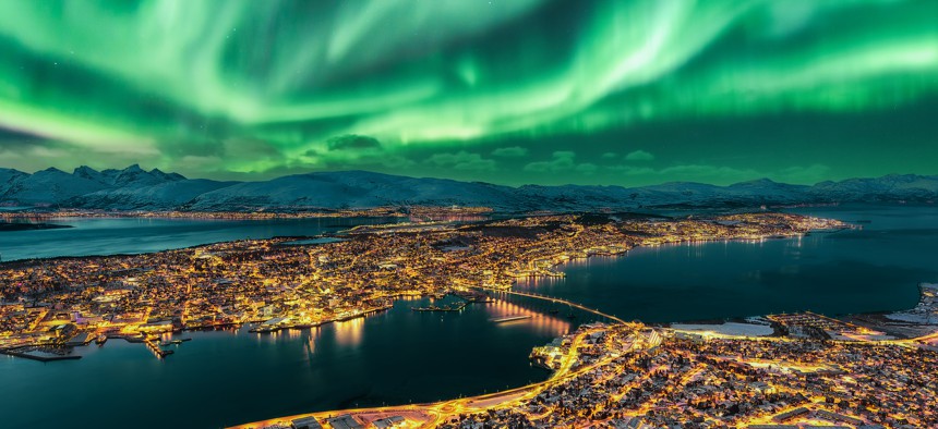 Aurora Borealis dancing over Tromso Urban Skyline. Tromso Panorama and winter night landscape with Northern Lights (Aurora Polaris) on a cold night, with starry sky, polar lights and snowcapped mountains on Kvaloya Island in the background. Cityscape with Brightly Street Light of the Arctic Capital, Northern Norway