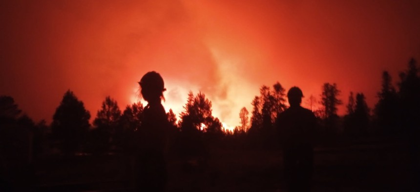 In this handout image provided by Grand Canyon National Park, Two firefighters stand silhouetted against an intense orange-red sky as the Dragon Bravo Fire burns through a forested area at night, with flames and smoke rising in the background at Grand Canyon National Park on July 12, 2025.