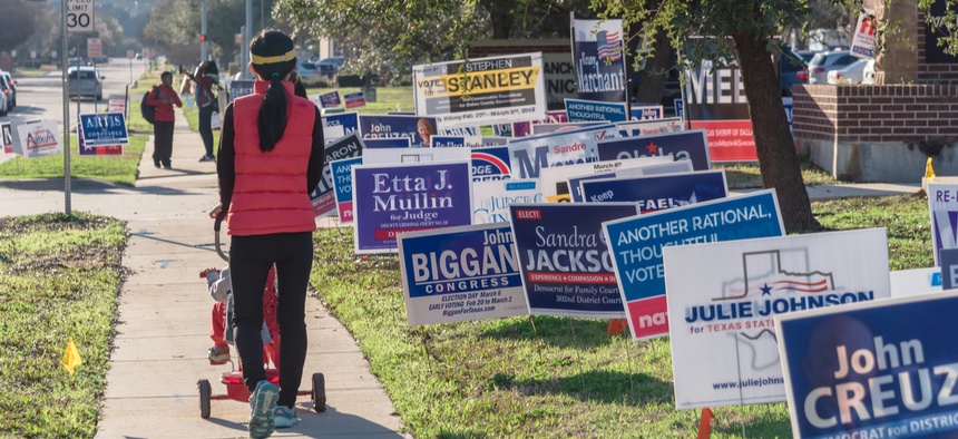 A mother pushes her child in a stroller through a residential area littered with campaign signs.